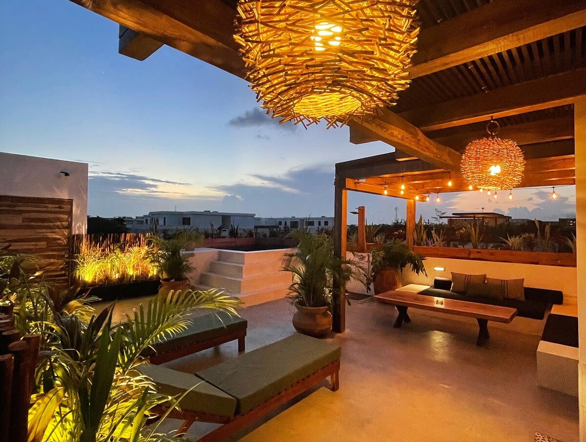 An inviting outdoor space is illuminated by warm lighting from unique fixtures. Comfortable seating arrangements are positioned among lush greenery, creating a tranquil atmosphere. Steps lead to a lower level, enhancing the layered design of the private rooftop terrace.