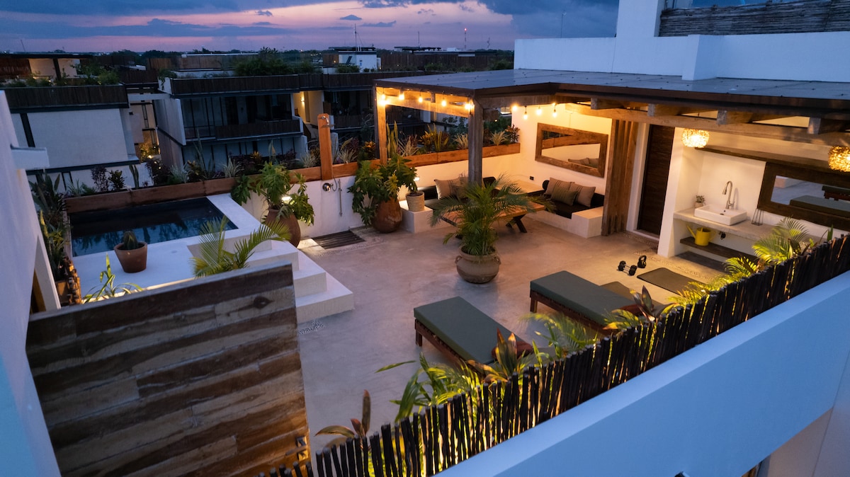A private rooftop terrace is illuminated by soft lighting, featuring a plunge pool and various seating areas with lounge chairs. Tropical plants provide natural privacy, and outdoor workout equipment is positioned strategically throughout the space, offering a serene ambiance for relaxation or exercise.