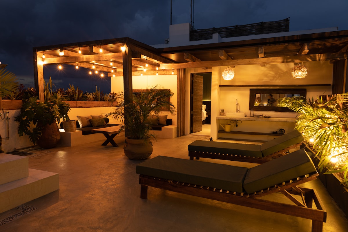 A spacious private rooftop terrace is illuminated by warm string lights, creating a serene ambiance. Comfortable lounge chairs are positioned for relaxation, surrounded by lush greenery. A small table and seating area offer additional space for gathering. The setting emphasizes a tranquil outdoor living experience.