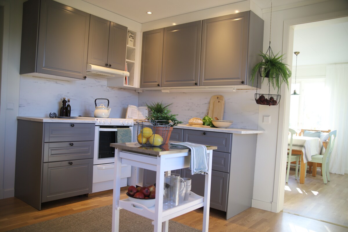 The kitchen features modern grey cabinetry and a white countertop. A stove and oven are visible, alongside a kettle and kitchen tools. Fresh fruits and greenery add a touch of nature. A dining area can be seen in the adjacent space, illuminated by natural light.
