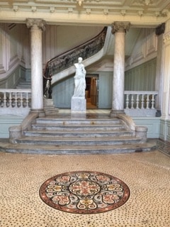 A grand entrance hall features a sweeping staircase adorned with a decorative railing. A statue stands at the top of the steps, while an intricate mosaic pattern lays on the floor. Elegant columns frame the space, accentuating the historic architectural details.