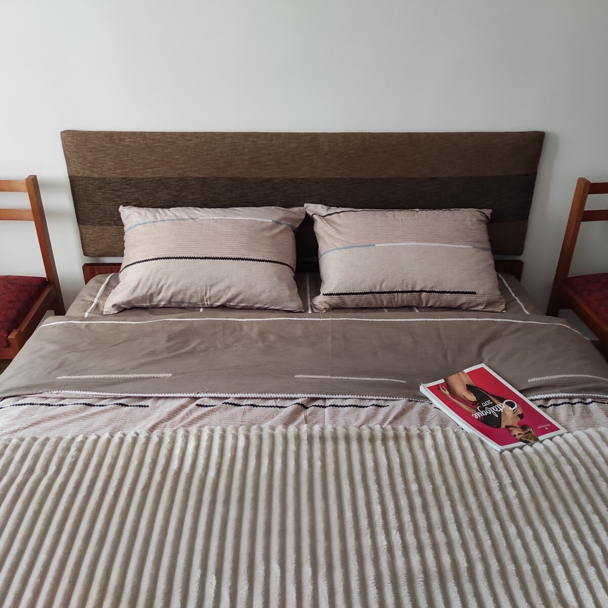 A cozy sleeping area features a spacious bed with neutral-toned bedding and two decorative pillows. A soft throw blanket is draped across the bed. A magazine rests on the bedspread, inviting relaxation. Two wooden chairs with red cushions are positioned beside the bed.