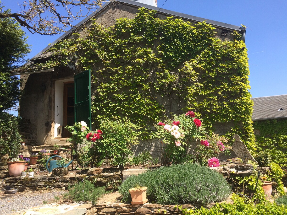 A charming exterior of a stone building is enveloped by lush greenery. Bright flowers in various colors add vibrancy to the garden, while a series of steps lead to the entrance, framed by large green doors. The roof is partially visible against a clear blue sky.