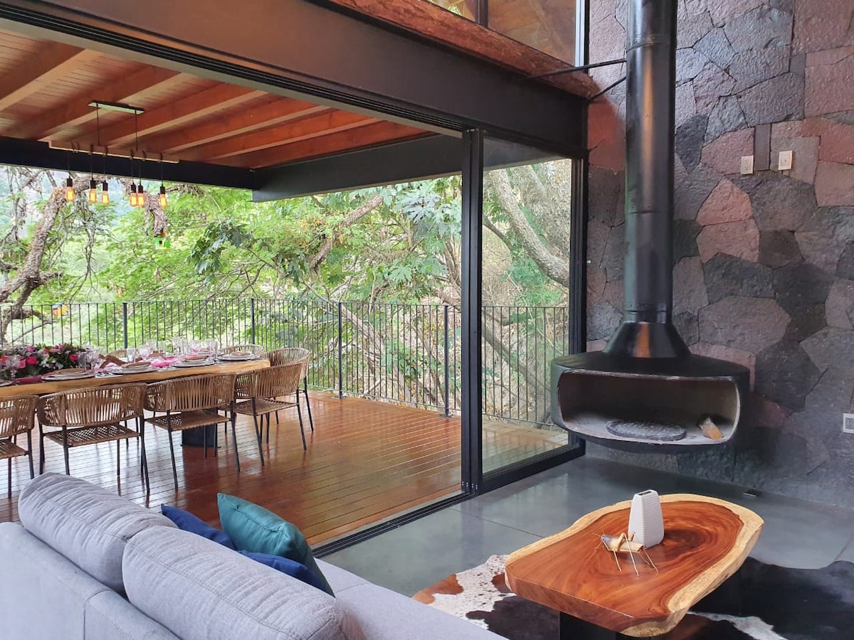 A cozy living area features a modern fireplace set against a stone wall, with large glass doors opening to an outdoor dining space. Natural light floods the interior, highlighting a wooden coffee table and a sectional sofa adorned with colored cushions.