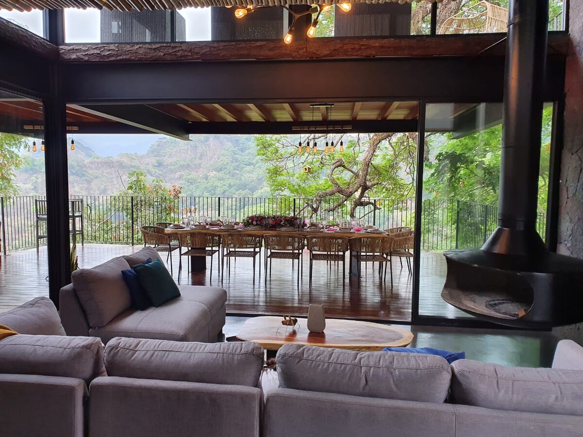 A spacious living area features a comfortable seating arrangement with light-colored sofas and a coffee table. Large glass doors offer an unobstructed view of the lush surroundings. A dining area with a long table is visible in the background, enhancing the connection to nature.