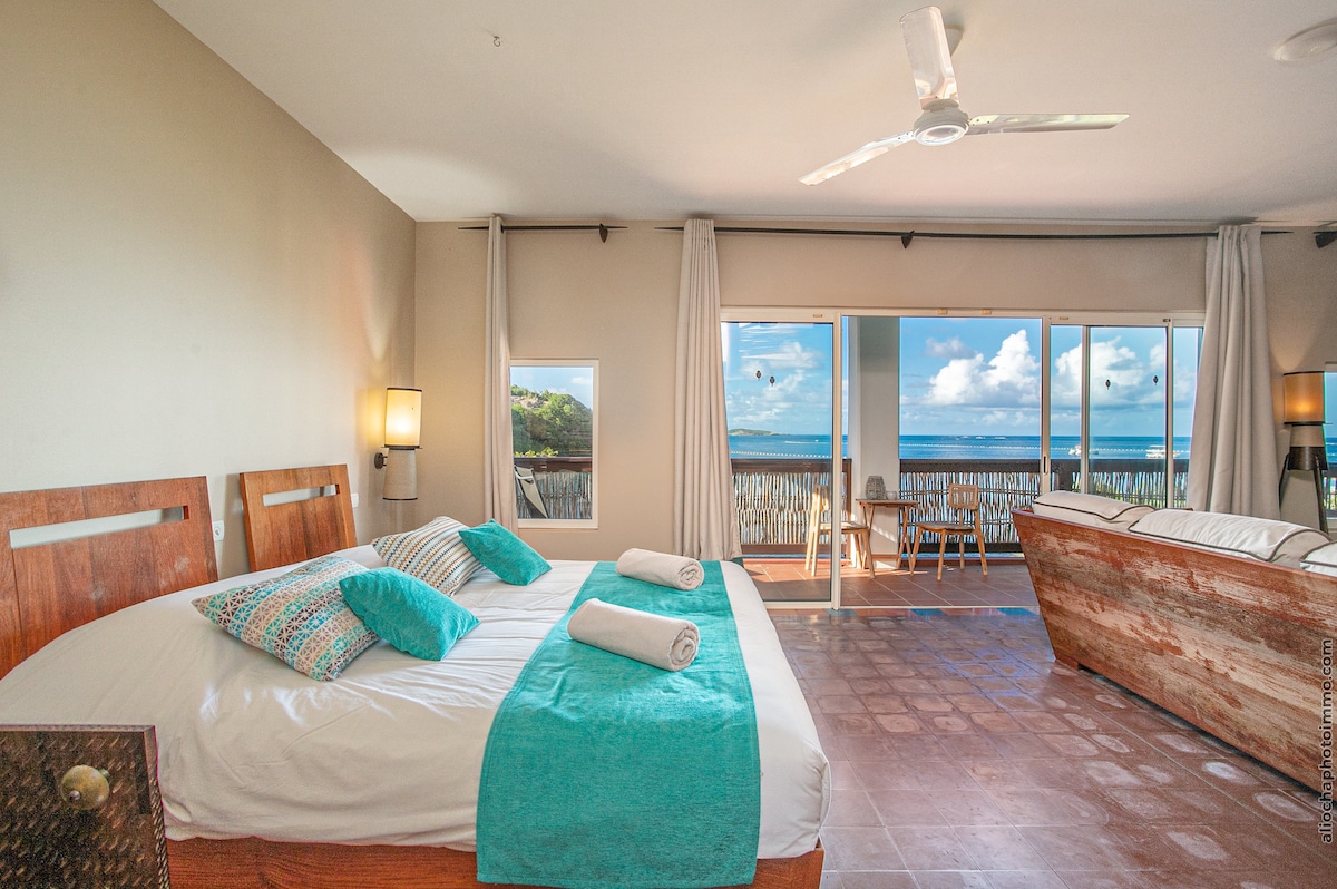 A spacious sleeping area displays a large bed with folded towels and decorative pillows. Large windows provide views of the sea, while natural light fills the space. A ceiling fan circulates air, and a seating area with wooden chairs complements the room.