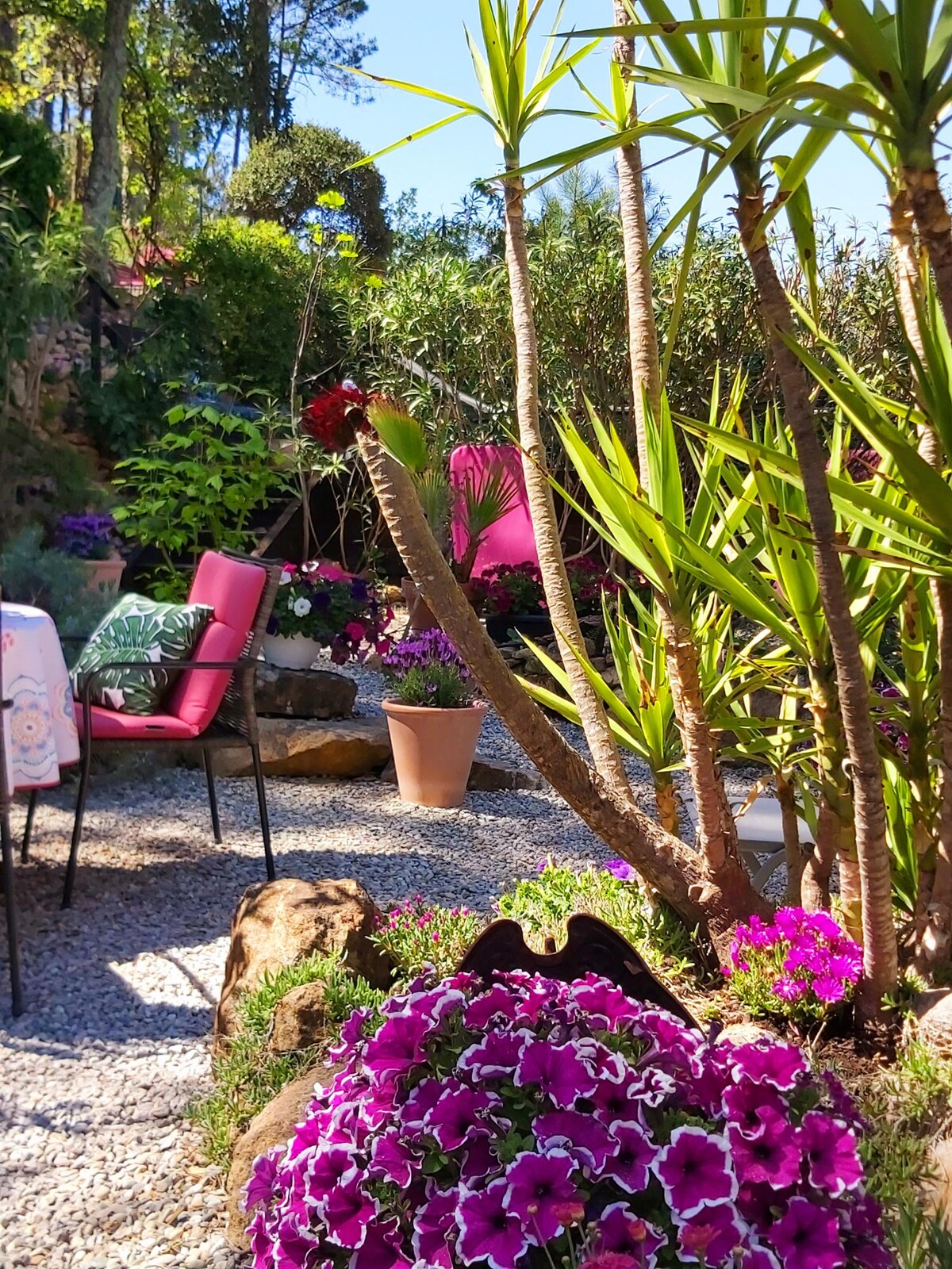 A lush private garden is depicted, featuring vibrant flower beds filled with pink and purple blossoms among greenery. A small, natural stone pathway leads to a seating area with two pink chairs, surrounded by potted plants and mature trees, creating a tranquil outdoor setting.
