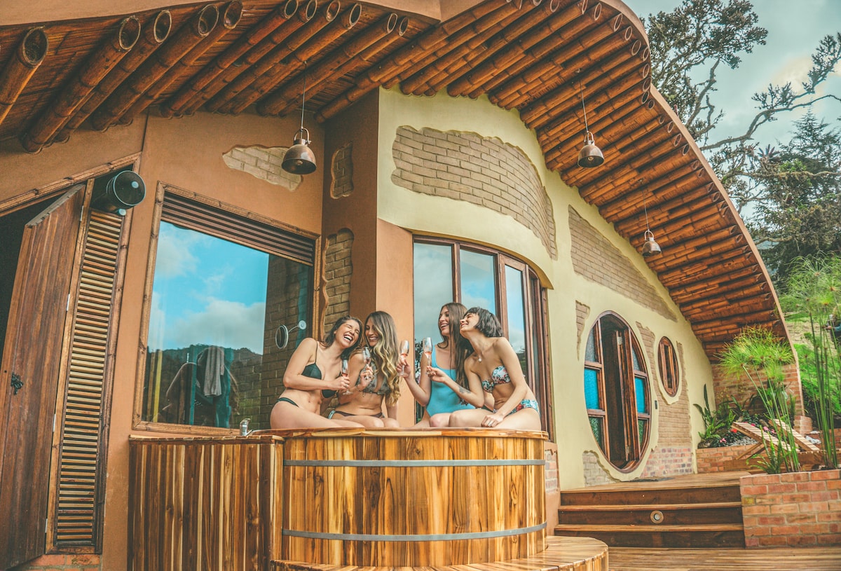 A unique house with natural stone walls and a curved roof is featured. A wooden deck surrounds a hot tub, inviting guests to relax while enjoying views of the surrounding nature. Large windows allow natural light to fill the interior space.