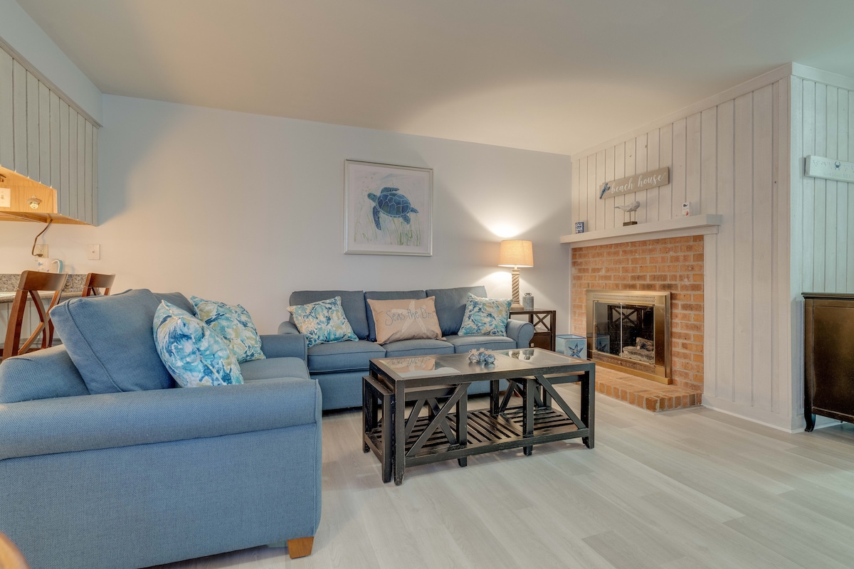 A comfortable living area features a spacious blue sectional sofa adorned with decorative pillows. A wooden coffee table sits at the center, and a fireplace is nestled in the corner, framed by a light-colored wall with paneling. Soft lighting creates a relaxed ambiance.