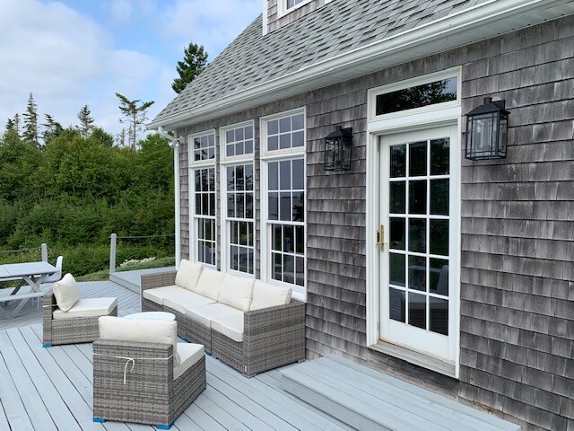 The outdoor seating area features a comfortable wicker sofa and chairs arranged on a spacious deck. Glass doors lead into the house, and lantern-style wall sconces flank the entrance. The surrounding greenery provides a natural backdrop for relaxation.