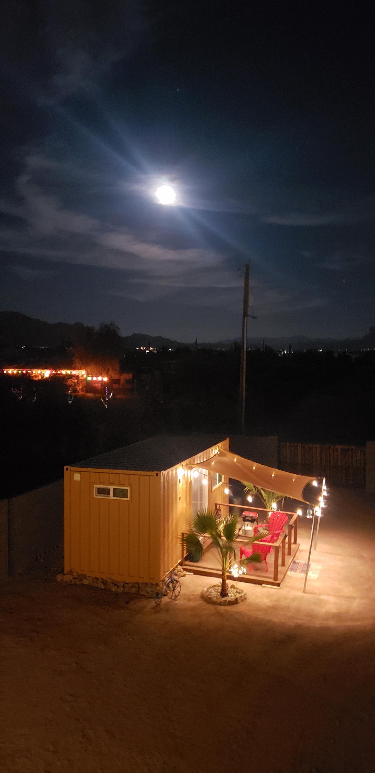 A mini home is illuminated under a bright moon, with outdoor string lights adding warmth to the space. A small deck features a table and chairs, surrounded by desert terrain. The distant mountains create a serene backdrop against the night sky.