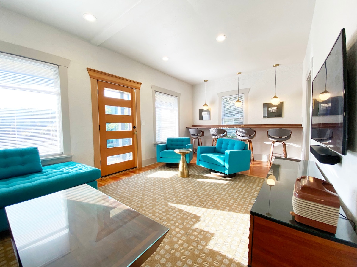 A mid-century modern living area is highlighted by two turquoise sofas and a glass coffee table. Warm light filters through the windows, creating a bright ambiance. A bar area with high stools is positioned against the wall, enhancing the stylish decor.