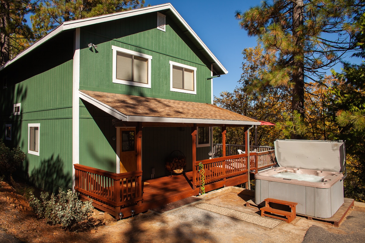 A green cabin is set against a clear blue sky, featuring a welcoming front porch with wooden railing. A spacious hot tub is situated on the porch, inviting relaxation. The surrounding area is filled with trees, enhancing the natural setting.