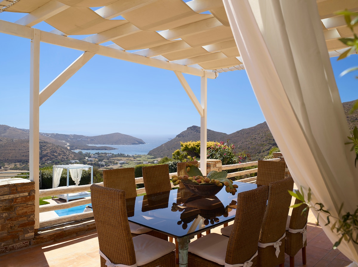 An outdoor dining area is presented under a shaded pergola, featuring a glass table surrounded by wicker chairs. A panoramic view of the Aegean Sea and hills is visible in the background, enhancing the space's inviting ambiance.