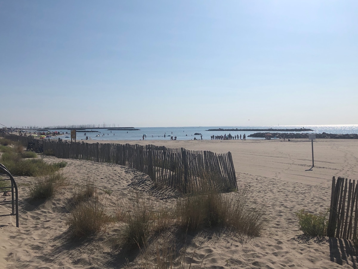 A sandy beach stretches out under a clear blue sky, with sunbathers and swimmers enjoying the water. Wooden fences line the beach, with patches of grass peeking through the sand. The calm sea reflects the sunlight, creating a serene coastal atmosphere.