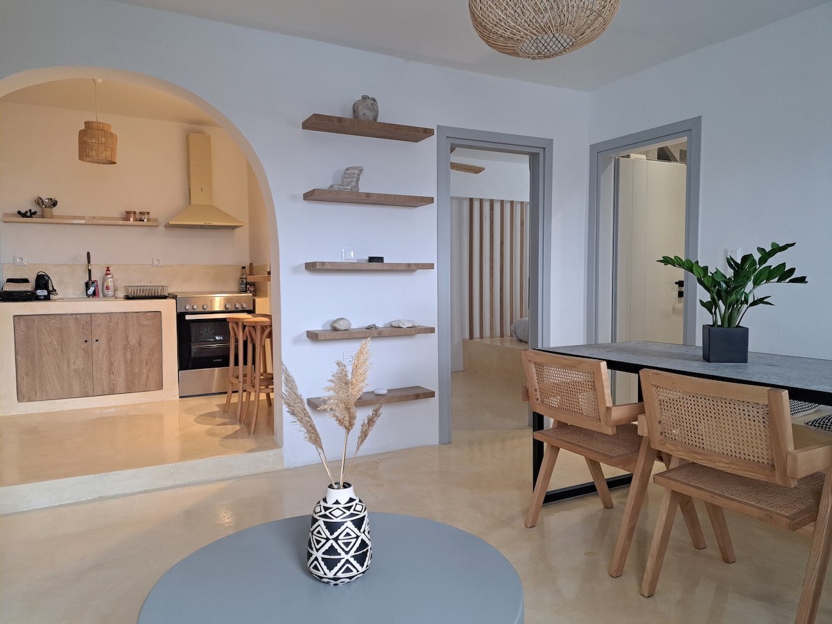 The open living area features a modern kitchen with wooden shelves and a stove, complemented by a dining table surrounded by two chairs. A decorative vase holds dried plants on a round coffee table, and the space is illuminated by natural light from the entryway.