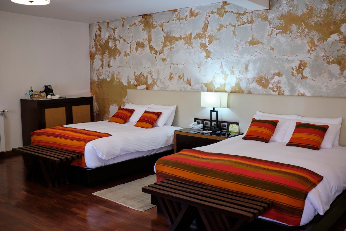 The room features two spacious queen beds, each adorned with colorful decorative blankets. A textured wall in warm tones adds character. A wooden bench is positioned at the foot of each bed. A bedside table holds a lamp and various amenities, enhancing functionality.