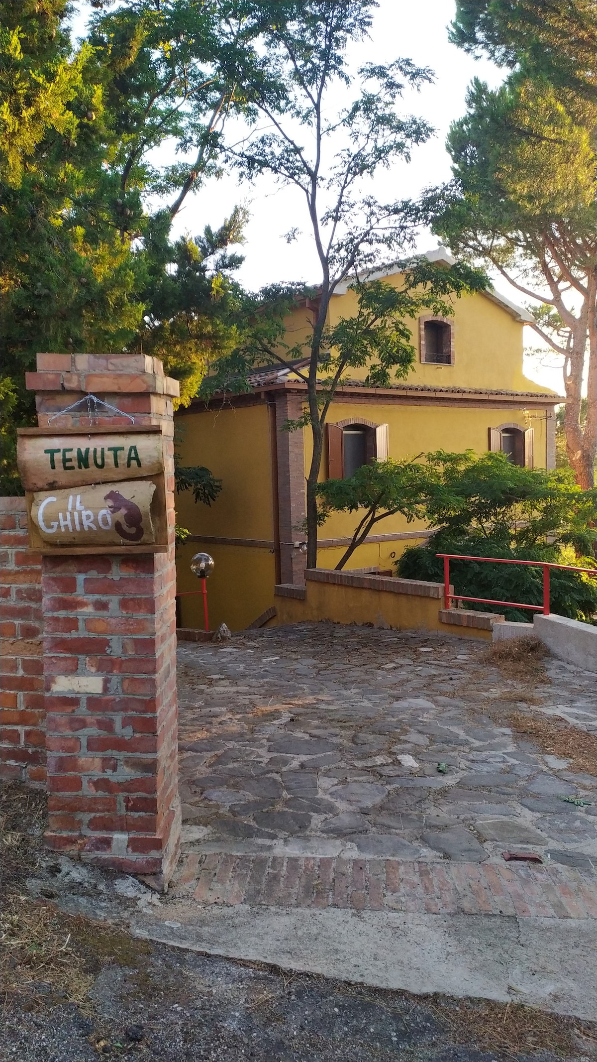 An inviting pathway leads to the entrance of Tenuta Il Ghiro, characterized by its warm yellow exterior and wooden windows. Surrounding trees provide natural shade, enhancing the tranquil setting. A stone path, complemented by a rustic brick wall, guides visitors towards the property.