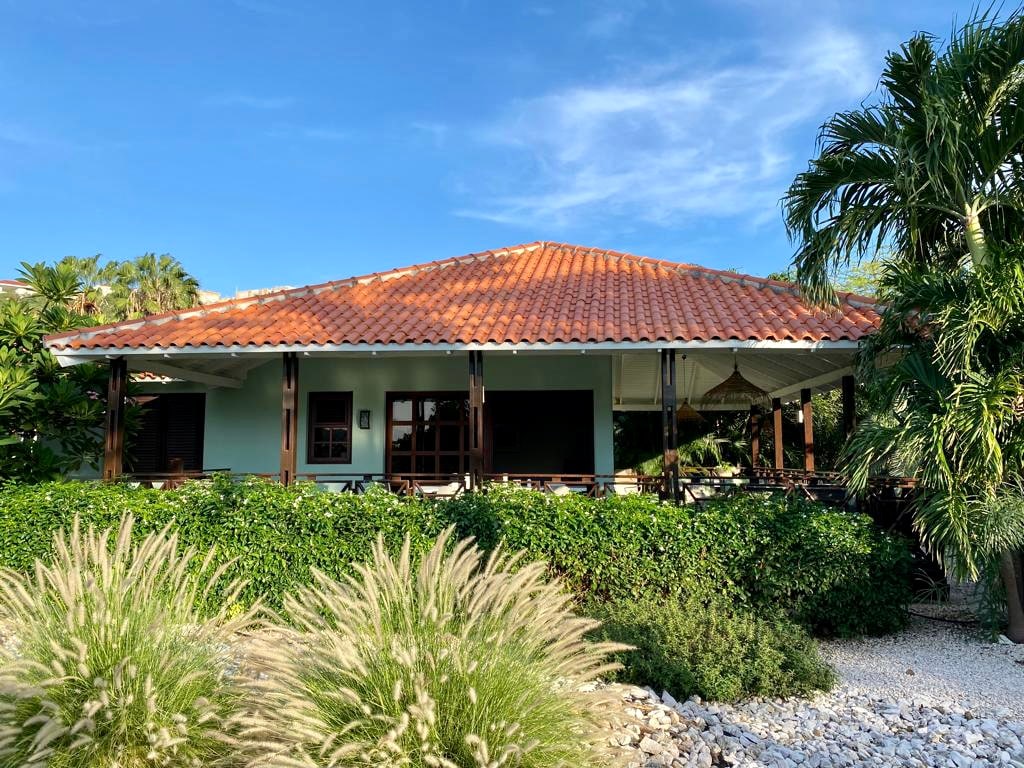 The villa features a charming facade with a terracotta roof and wooden accents. Lush greenery surrounds the property, creating a private atmosphere. The spacious terrace is partially shaded, offering a comfortable outdoor seating area to enjoy the views of the tropical surroundings.