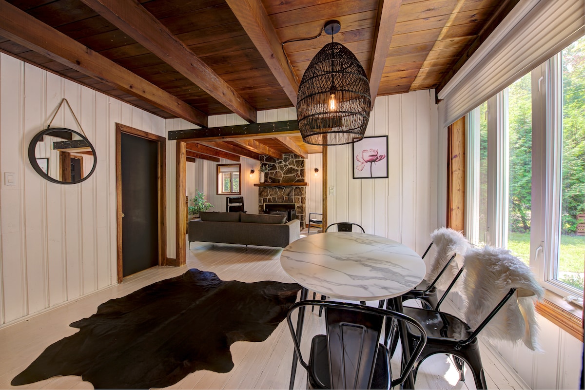 A dining area features a round marble table surrounded by black chairs, with a decorative cowhide rug beneath. Natural light enters through large windows, illuminating rustic wood beams and a cozy living space in the background, complete with an inviting stone fireplace.