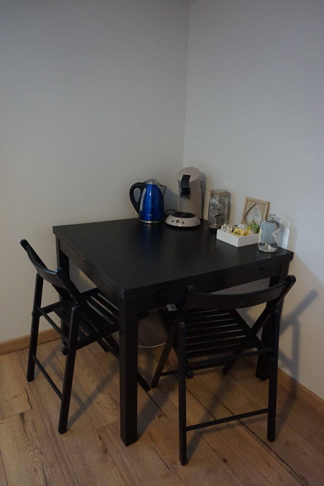 A compact corner dining area is shown, featuring a black table with two matching chairs. A kettle and a coffee machine are placed on the table, along with a small decorative arrangement. Natural wood flooring complements the space.
