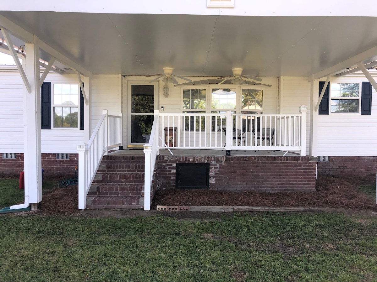 A covered porch is presented with white railings and ceiling fans, offering a shaded area. Large windows are visible, allowing natural light to fill the space. Brick steps lead up to the entrance, with a grassy area surrounding the porch.