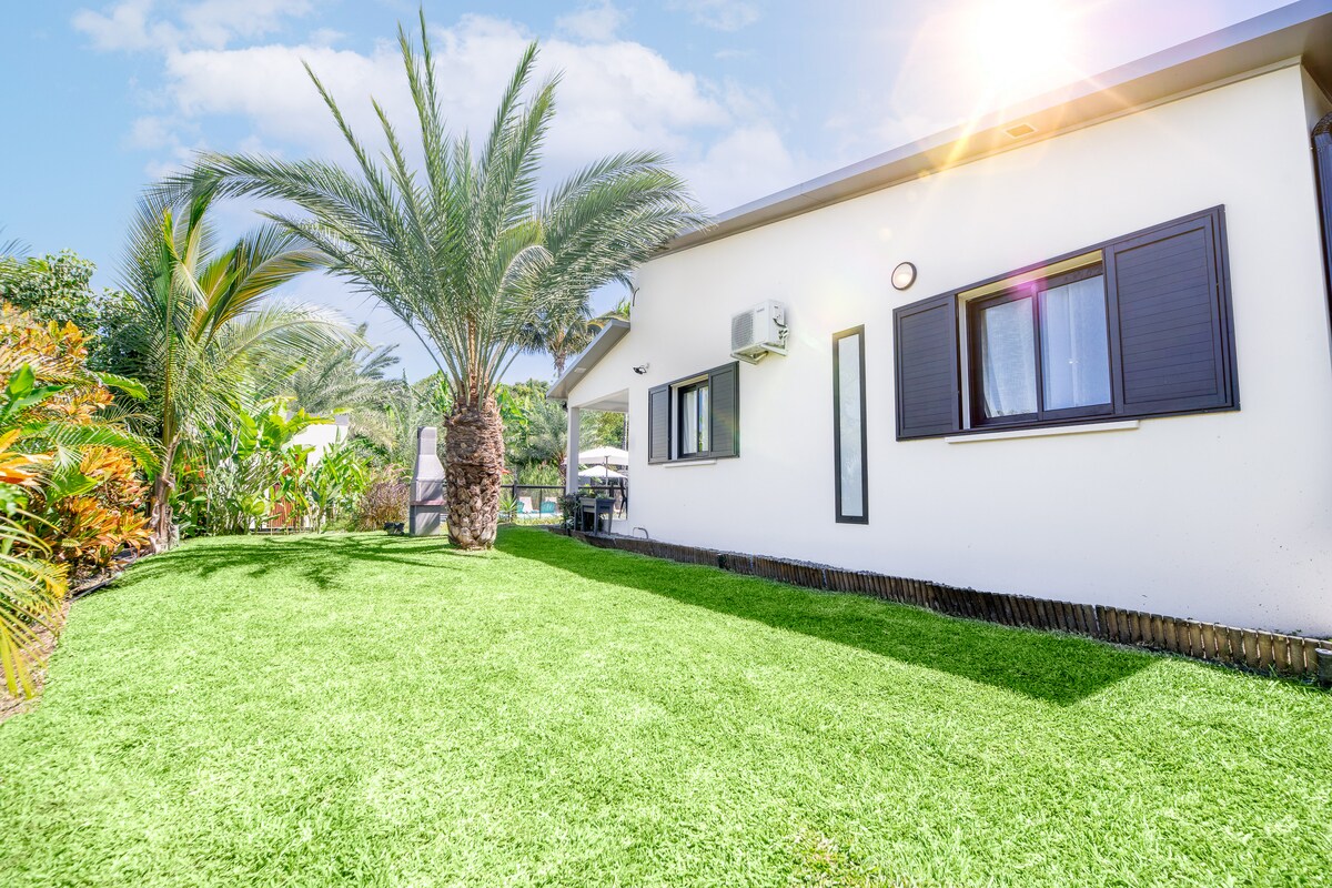 A spacious lawn is presented, bordered by lush greenery and palm trees. The exterior of the lodging features large windows with dark shutters, providing natural light. A clear blue sky complements the tranquil setting of this inviting outdoor space.