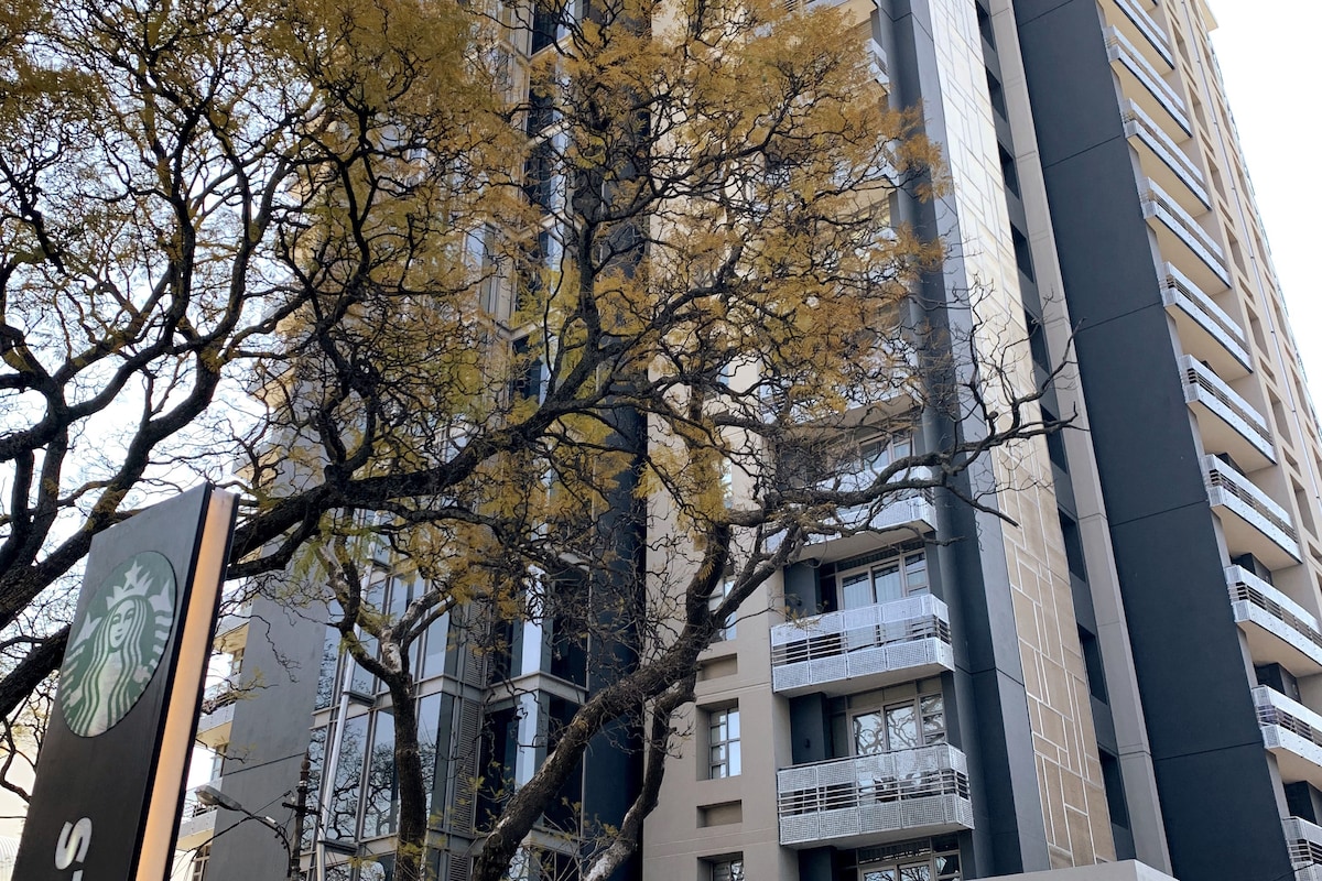 A modern high-rise building stands prominently against a clear sky, framed by the skeletal branches of trees. The distinctive Starbucks sign is visible in the foreground, contributing to the urban setting.