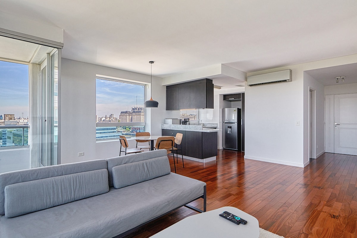 A modern living space is highlighted by large windows offering views of the city skyline. The area features a light grey sofa and a minimalist coffee table, with a dining space set for two. The open-concept kitchen includes sleek cabinetry and stainless steel appliances.