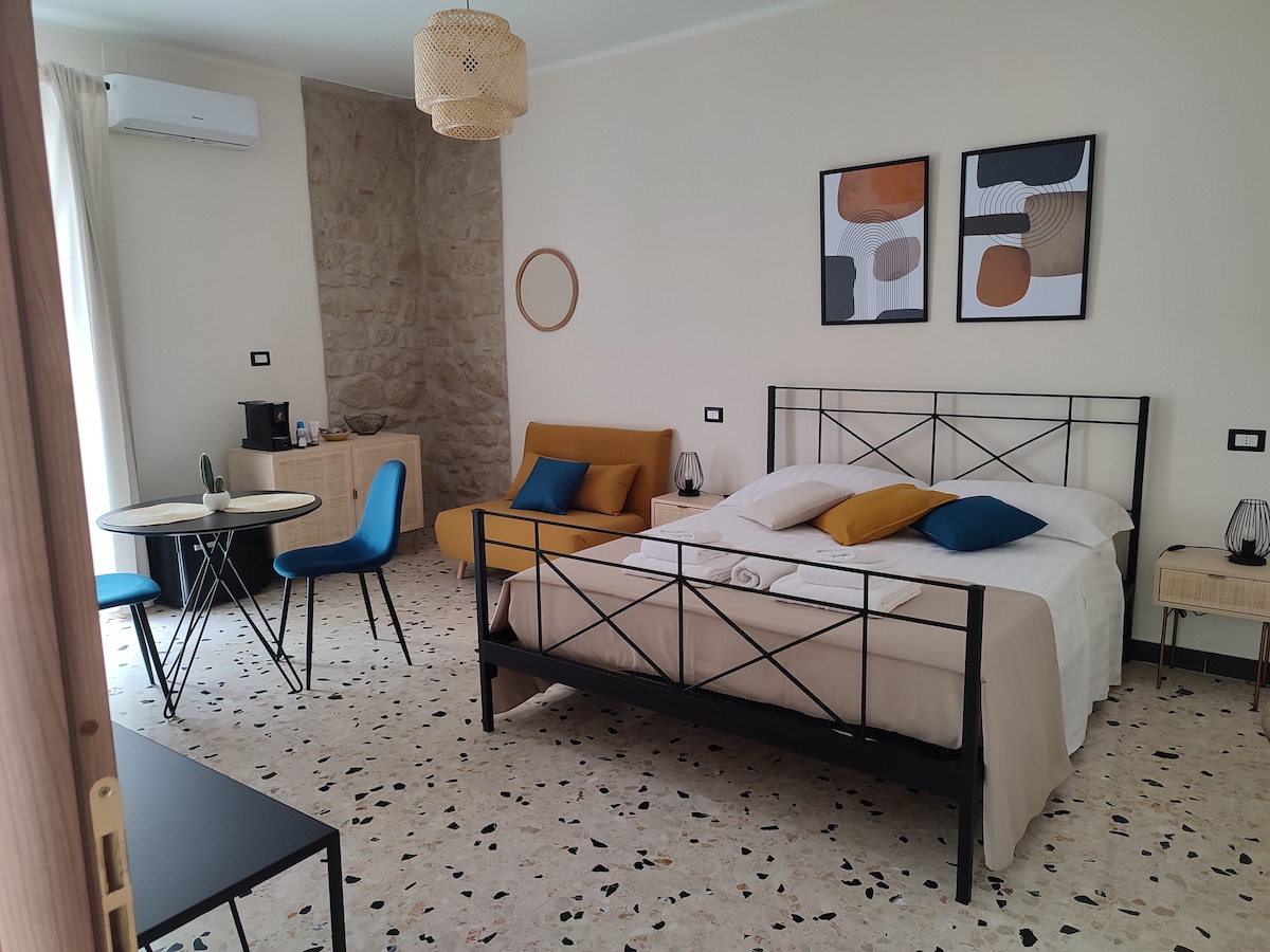 A spacious room features a large bed with a neutral-toned duvet, complemented by colorful pillows. A seating area includes a single sofa and a small round table with two chairs. Decorative wall art and a light fixture enhance the ambiance, while a large window introduces natural light.
