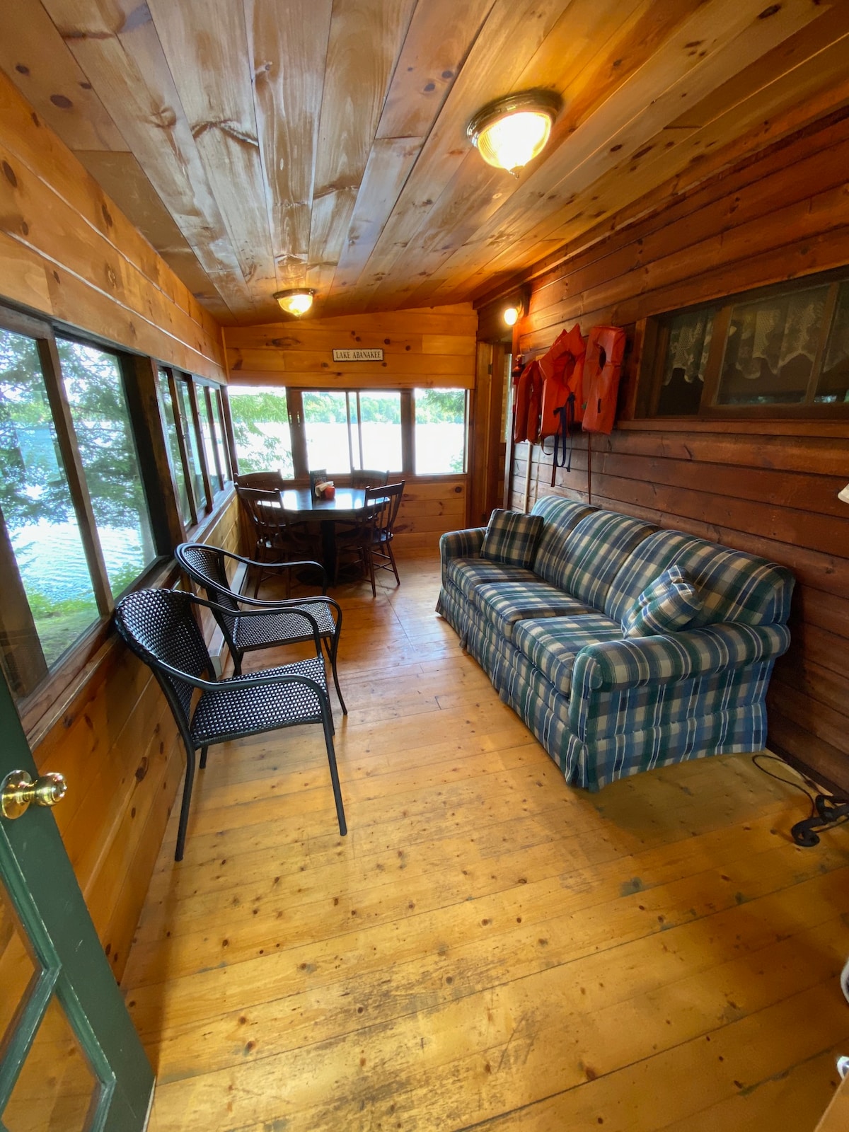 A screened-in porch features wooden walls and a sunlit view of the lake. A plaid couch provides seating, accompanied by four black wicker chairs. A small table sits nearby, creating a cozy area for relaxation or conversation.