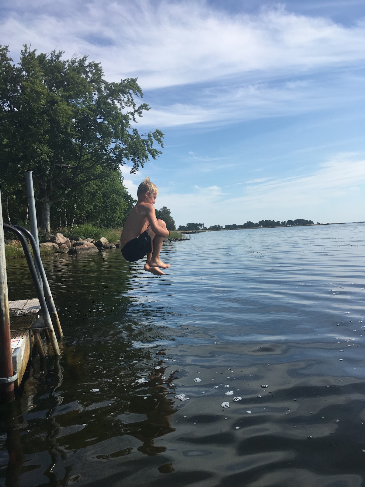 A child is captured in mid-air, preparing to jump into a calm body of water. Sunlight brightens the scene, reflecting off the water's surface, while trees line the shore in the background. The gentle ripples suggest a serene environment.