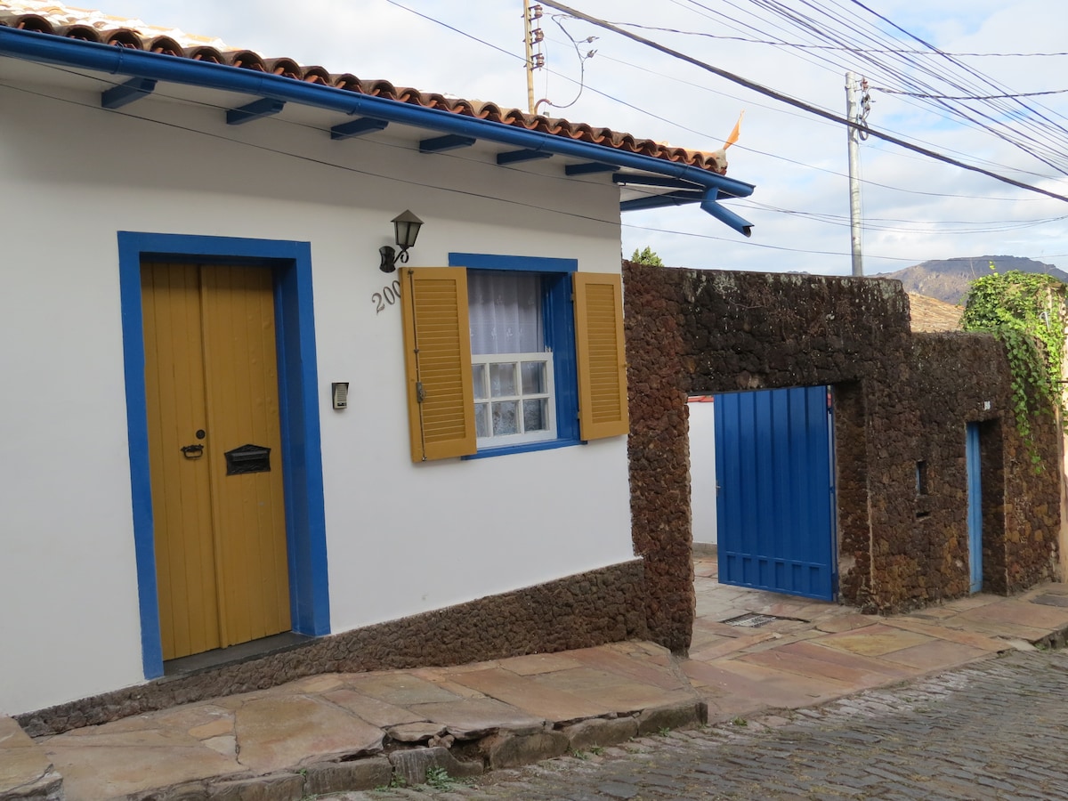 The exterior of a charming house is displayed, featuring a yellow door with a lantern and blue shutters framing the windows. A stone wall and gate provide access, while the cobblestone path adds texture. Natural greenery climbs along the wall, enhancing the inviting appearance.