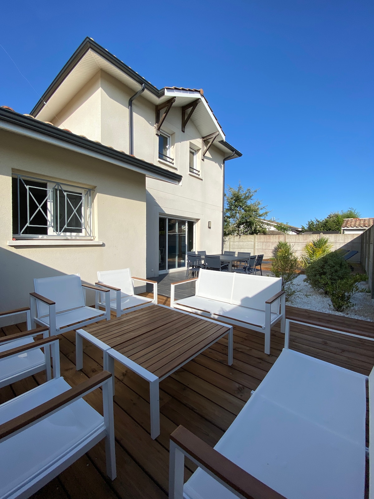 An outdoor seating area is featured, with a wooden table surrounded by six white chairs, set on a wooden deck. A spacious sliding door leads to the interior. The clear blue sky provides a bright ambiance, while green foliage adds a touch of nature.