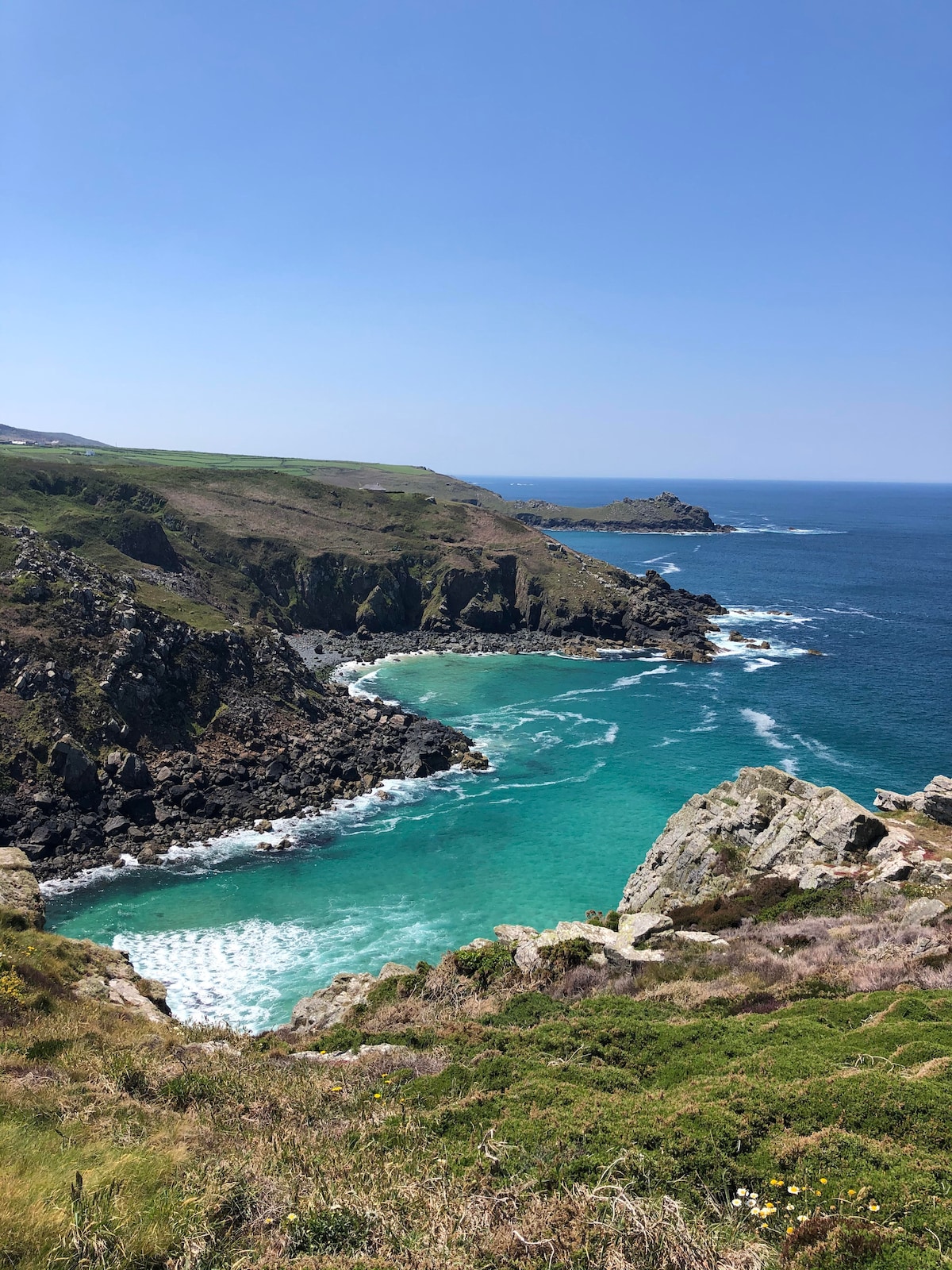 A scenic view of the rugged Cornish coastline is captured, showcasing lush green cliffs contrasting with vibrant turquoise waters. Gentle waves lap against the rocky shore, while distant land formations create a horizon under a clear blue sky.