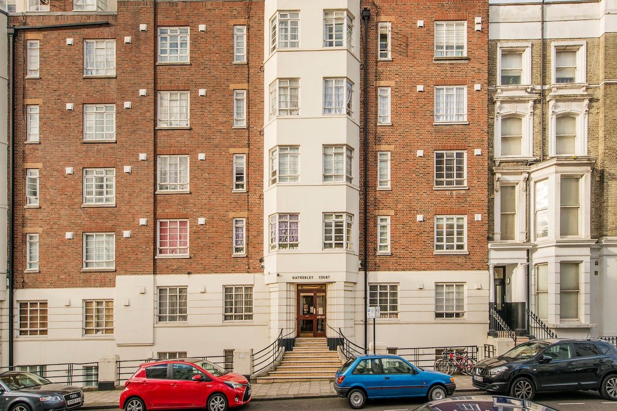 A well-maintained brick building with a classic facade features multiple windows in various styles, allowing natural light to fill the interiors. The entrance is highlighted by a set of stairs leading to the doorway, flanked by parked cars along the street.