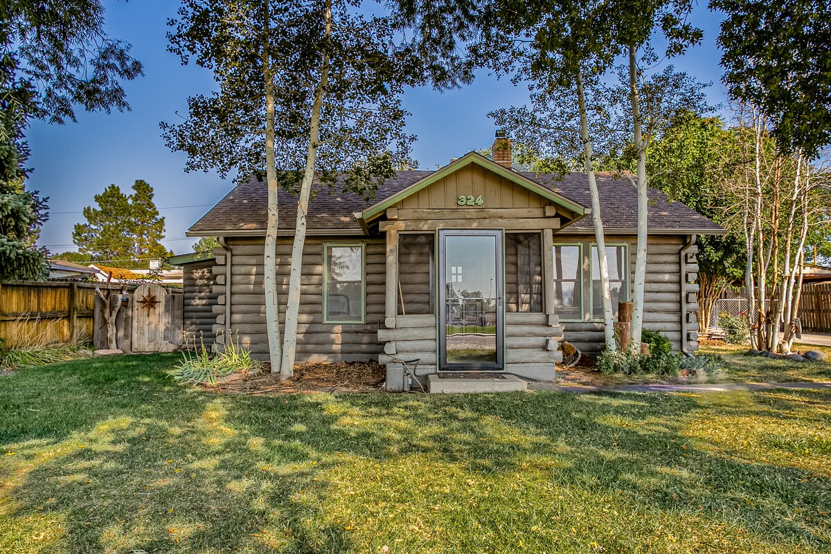 The charming exterior of a remodeled 1940's cabin is presented, showcasing a log cabin style with a welcoming front porch. Surrounding trees provide shade, and a spacious lawn enhances the outdoor appeal. A glass front door offers a glimpse into the cozy interior.