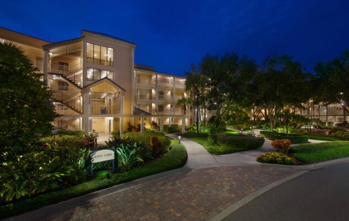The exterior of the building is illuminated at night, showcasing a blend of architectural features and lush landscaping. Pathways meander through well-maintained gardens, and soft lighting enhances the inviting atmosphere of the surroundings.
