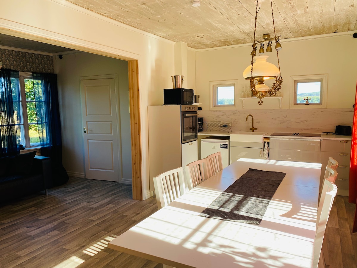 A newly renovated kitchen area is visible, featuring sleek cabinetry and modern appliances. A large dining table with white chairs is positioned in the center, illuminated by natural light streaming through the windows. Counters showcase a blend of functionality and style.
