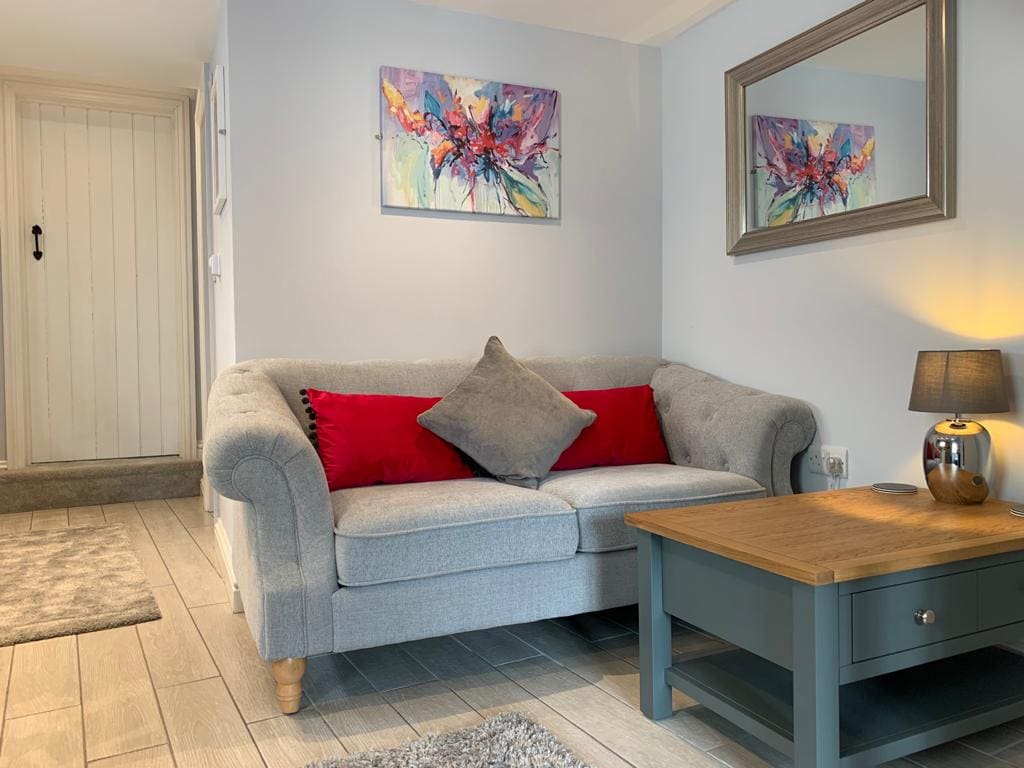 A light-filled living area features a gray sofa adorned with red and gray cushions. A wooden coffee table rests on a soft rug, while vibrant artwork enhances the white walls. A mirror reflects the space, and a small lamp provides a warm glow.