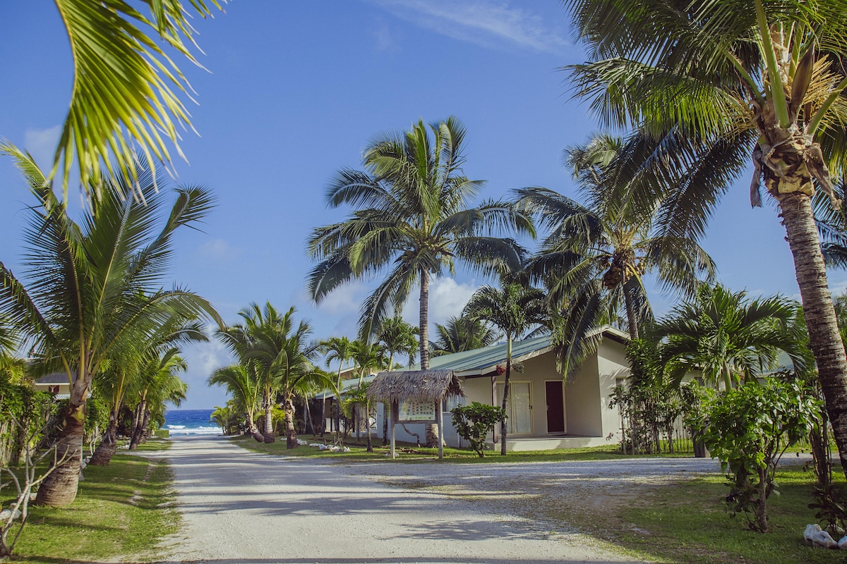 A sandy pathway lined with palm trees leads to the beach, creating a serene atmosphere. On either side, garden villas are visible, each surrounded by lush greenery, under a clear blue sky with a hint of ocean in the distance.