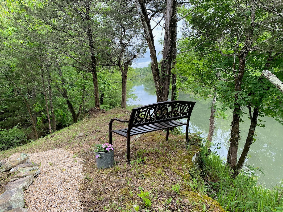 A black metal bench is positioned under leafy trees overlooking the Little River. A small planter with pink flowers sits beside the bench, enhancing the serene riverside setting. The area is surrounded by lush greenery, creating a peaceful and natural atmosphere.