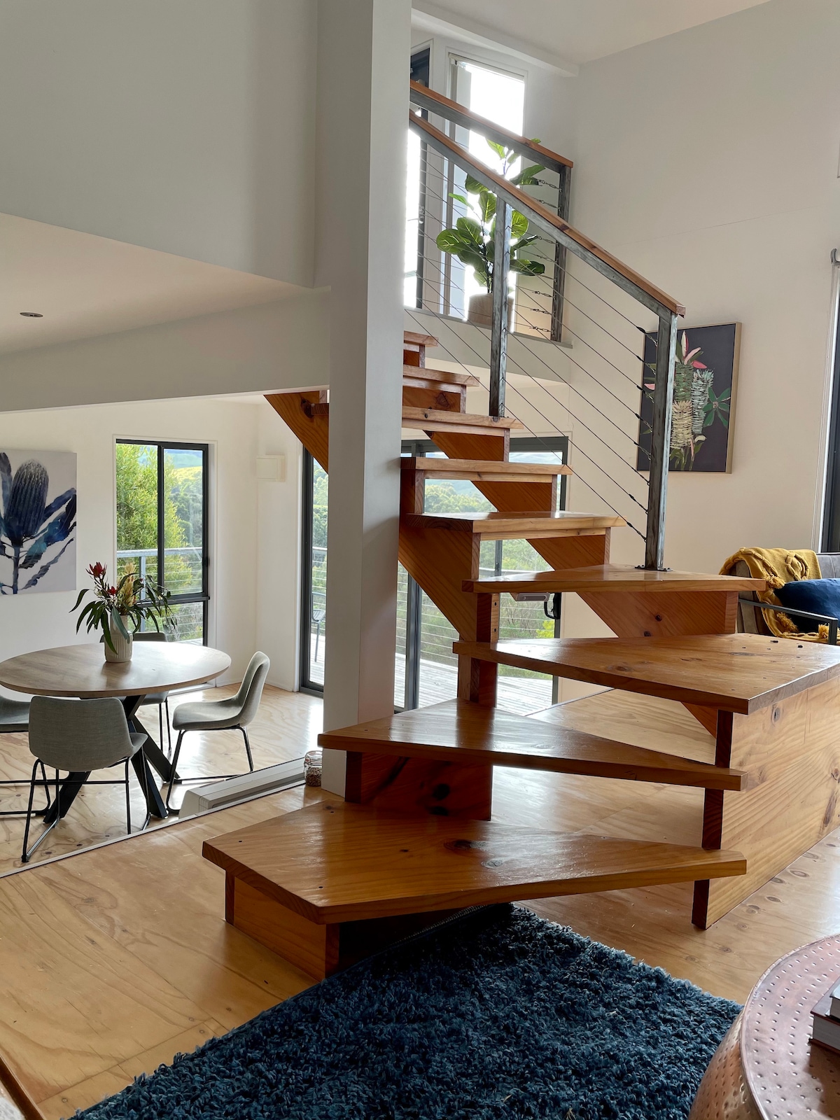 A spiral wooden staircase leads from the lower floor to the upper level, showcasing a modern design. A round dining table is positioned near a large window, allowing natural light to fill the space. Decorative plants and artwork adorn the walls, enhancing the natural setting.