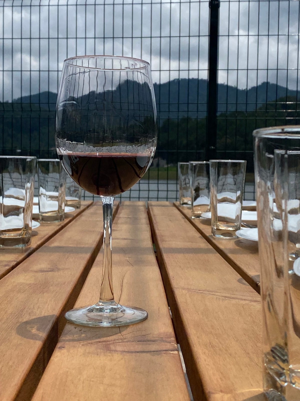 A glass of red wine is positioned on a wooden table, surrounded by clear glasses. The background features a view of distant mountains under a cloudy sky. The table is set, suggesting a welcoming atmosphere for gatherings.