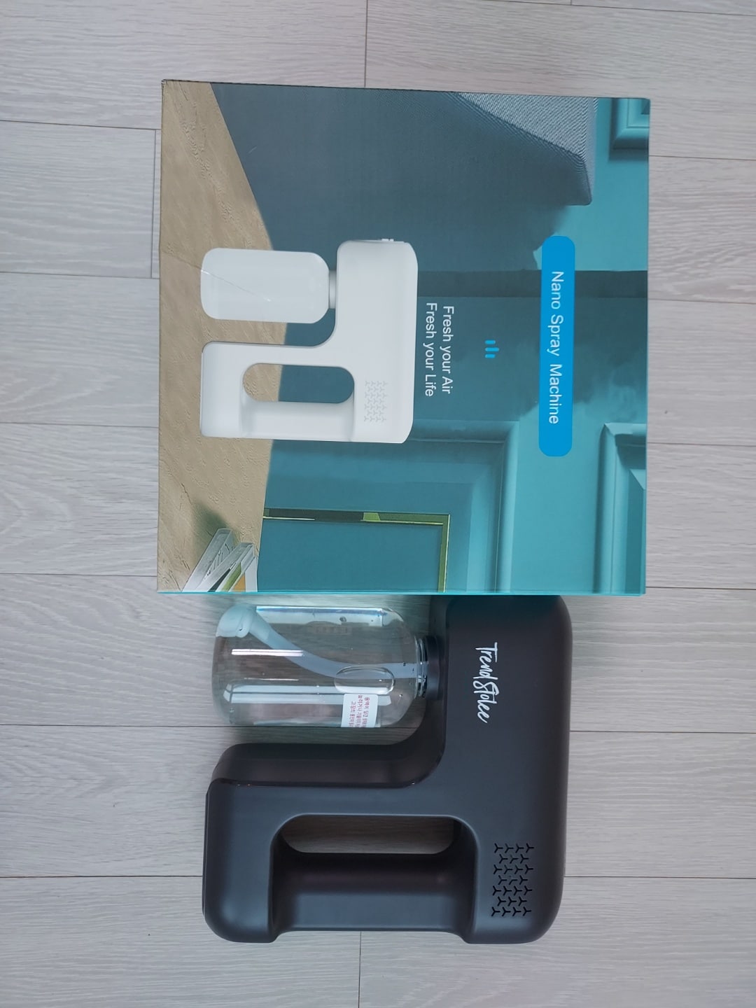 An air spray machine is displayed beside its packaging. The device is predominantly white with a black handle and a clear water container. The box features a modern design and product information, contrasting with the light wooden flooring beneath.