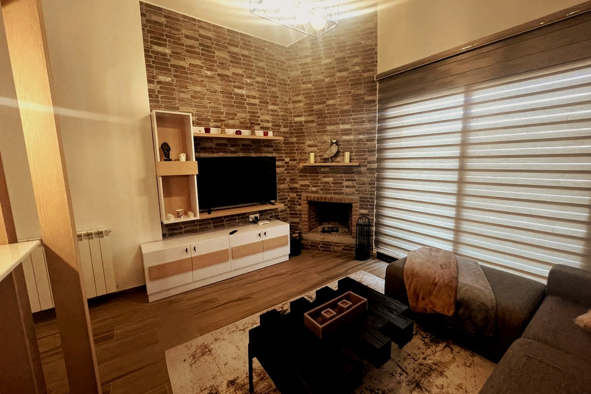 A modern living area is showcased, featuring a comfortable sofa and a sleek coffee table. A television is mounted on a stylish cabinetry, complemented by a decorative fireplace. Natural light filters through layered window treatments, enhancing the warm tones of the stone accent wall.