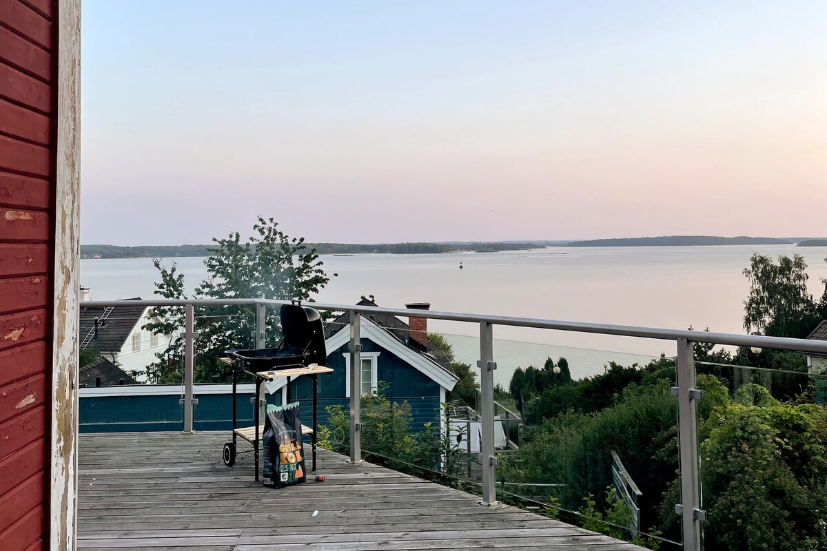 A spacious deck offers an unobstructed view of the calm water, reflecting a soft sunset hue. A barbecue grill is positioned on the wooden floor, surrounded by lush greenery and distant rooftops, contributing to a peaceful and serene outdoor setting.