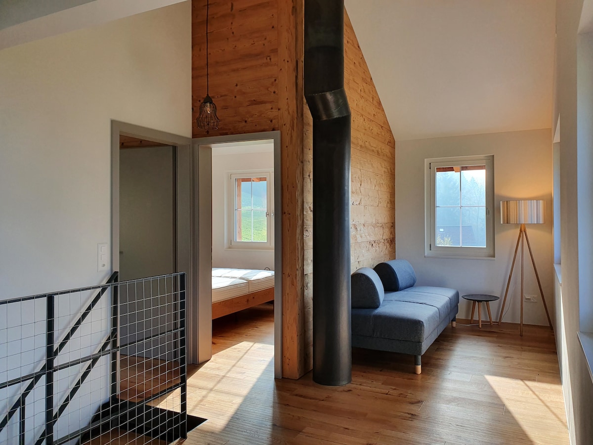 A spacious living area features a gray sofa, a tall lamp beside it, and a wooden side table. Light filters through two windows, highlighting the natural wood wall and the sleek, modern staircase. An adjoining bedroom is subtly visible through an open doorway.
