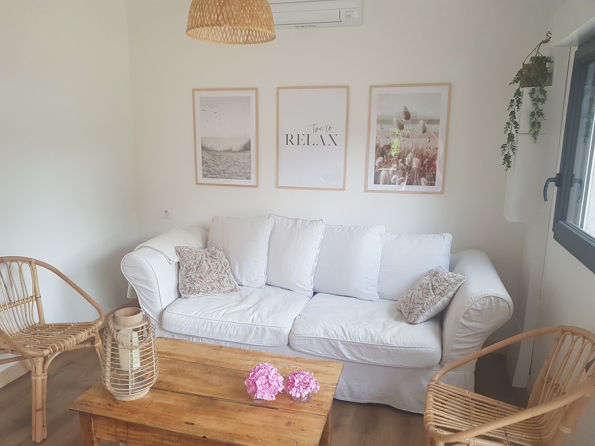 A light-filled seating area is furnished with a white sofa and natural wood coffee table. Two woven chairs offer additional seating options. The walls feature three framed art pieces, and a small plant hangs near the window, adding a touch of greenery to the space.