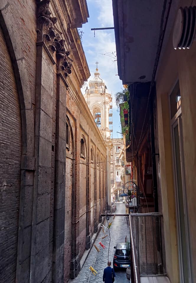 A narrow street is seen from a balcony, flanked by historic stone buildings. In the distance, a bell tower rises above the architecture, reaching towards a blue sky. The street is paved and lined with scattered debris, indicating previous activity.